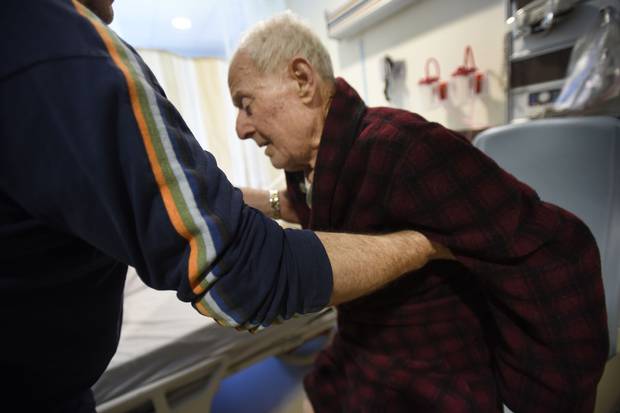 Tom Spanidis, left, helps his father out of his chair. The younger Mr. Spanidis says that when he refused to take his father home, hospital officials promised to start charging him $1,100 a day to stay.