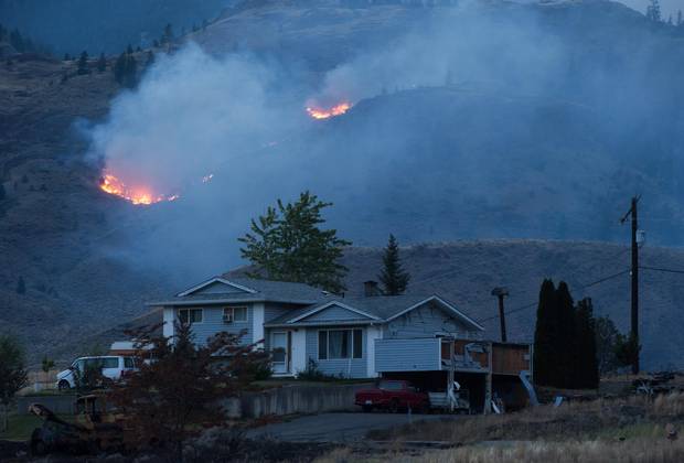 A wildfire burns on a mountain in the distance behind a house that remains standing on the Ashcroft First Nation, near Ashcroft, B.C., on Sunday July 9.