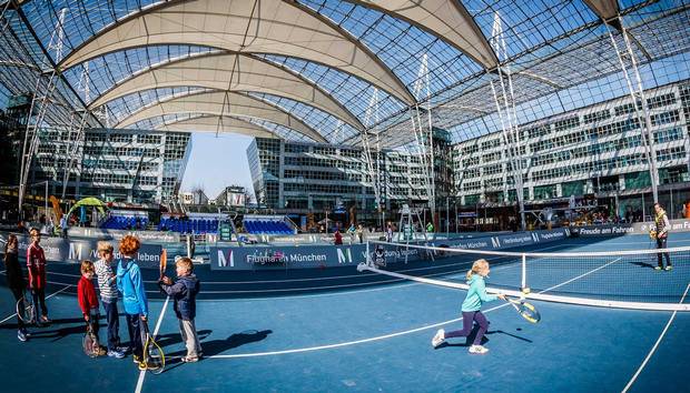 A large, covered outdoor space at Munich Airport features two tennis courts.