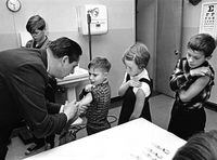 THAT OLD NEEDLE DOESN'T REALLY HURT... MUCH -- Measles vaccination for Toronto school children. Paul Cornier, middle, is more concerned, and Linda Dool watches with serious expression, as Dr. Michael Kapusta gives vaccinations at St. Teresa's  Separate School, New Toronto, December 13, 1965. New Toronto school children are being immunized in mass program against the disease.  Photo by James Lewcun / The Globe and Mail.Originally published December 14, 1965