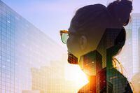 Business woman wearing sunglasses at sunset is looking at corporate office building. Double exposure photography