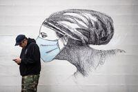 A man looks at his phone while standing near artwork of a woman wearing a protective face mask, on the side of a building in Vancouver, on Sunday, May 31, 2020. THE CANADIAN PRESS/Darryl Dyck