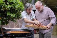 This image provided by Discovery shows Jose Andres holding a tray with ready cooked belly fat (torrezno) as Diego Guerrero sets it on a rice made in a paella pan in a scene from the Discovery + television series "Jose Andres and Family in Spain." (Xaume Olleros/Discovery via AP)