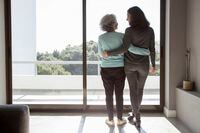 Rear view of happy mother and daughter standing embracing at window. Senior and mid adult women hugging and talking at home. Family relationships concept