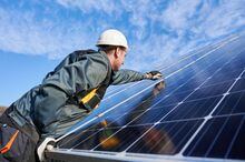 Side view snapshot of workman, wearing uniform, working gloves and helmet, setting a shiny new solar battery with help of hex key, blue sky on background. Green energy concept