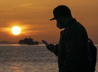 A man wearing a protective face mask to help prevent the spread of COVID-19 checks his phone as the sun sets in English Bay in Vancouver on April 5, 2021. Canada's existing mobile phone services and consumer groups will get a landmark ruling from the CRTC this afternoon. The regulatory ruling could shift some of the market power held by Rogers, Bell and Telus, which collectively have more than 90 per cent of the country's subscribers. THE CANADIAN PRESS/Jonathan Hayward