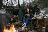 Dan Ebata (Left), Shelly Woodside (Middle), and Ryan Bellaire (Right) enjoy their morning coffee by the fire, warming up on a chilly -2°C day in the boreal forest of Canadian Shield near Ruth Lake in Killarney, Ontario. Shelly dries his boots by the fire, while Ryan sweetens his coffee with maple syrup. Captured on January 23rd, 2024."