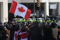 A person waves a Canadian flag as police on horseback and an armoured police vehicle are positioned in front of protesters, on the 22nd day of a protest against COVID-19 measures that has grown into a broader anti-government protest, in Ottawa, on Friday, Feb. 18, 2022. Lawyers for the federal attorney general are set to expand today on the legal rationale for the historic use of the Emergencies Act to dispel "Freedom Convoy" protesters early last year. THE CANADIAN PRESS/Justin Tang