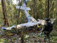 In this photo released by Colombia's Armed Forces Press Office, a soldier stands in front of the wreckage of a Cessna C206, Thursday, May 18, 2023, that crashed in the jungle of Solano in the Caqueta state of Colombia.  A search continues for four Indigenous children who may have survived the deadly plane crash in the Amazon jungle on May 1. On Tuesday, May 16, soldiers found the wreckage and the bodies of three adults, including the pilot and the children's mother. (Colombia's Armed Forces Press Office via AP)