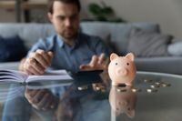 Close up of toy pink piggy bank and coins with young man using calculator and writing notes in background. Homeowner calculating savings, counting budget, doing finance control work